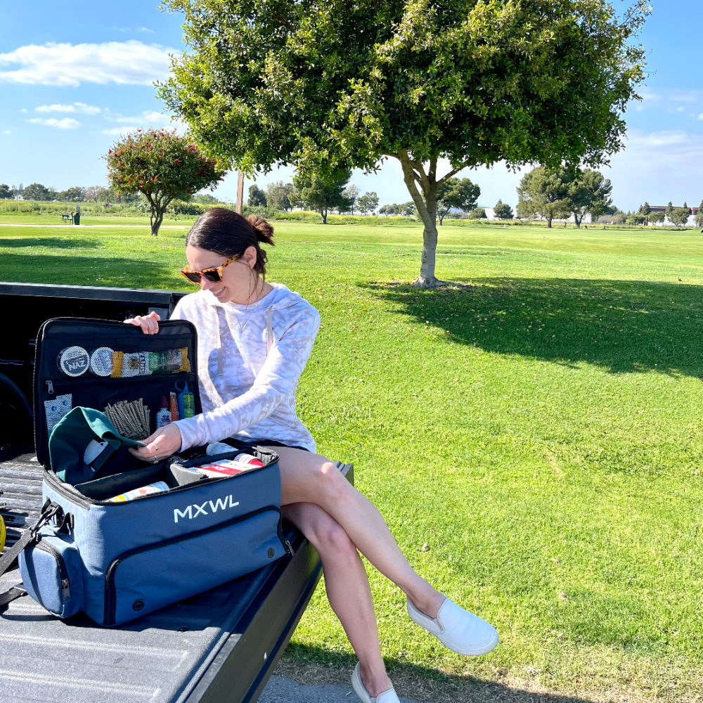Woman sitting on a truck bed with an open blue cooler labeled 'MXWL' in a park.